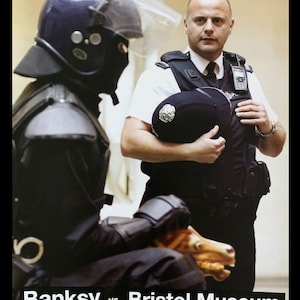 Op de afbeelding: Een zwartgeklede oproerpolitieagent staat naast een politieagent in uniform en houdt een zwarte helm vast. De tekst "Banksy vs Bristol Museum" is in zwart op een witte achtergrond gedrukt, met de data "June 13th - August 31st 2009" en "admission free" eronder gedrukt.