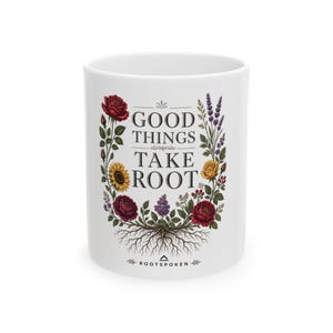 May include: A white ceramic mug featuring the black text GOOD THINGS TAKE ROOT centered between a colorful illustration of various flowers, leaves, and visible roots. The design includes red roses, a yellow sunflower, and purple lavender.