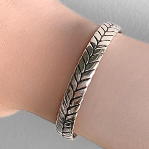 May include: Silver bracelet with a textured, woven pattern. The bracelet has a solid band design, with a pattern of overlapping, leaf-like shapes. The jewellery is worn on a wrist, highlighting the bracelet's details and shine.