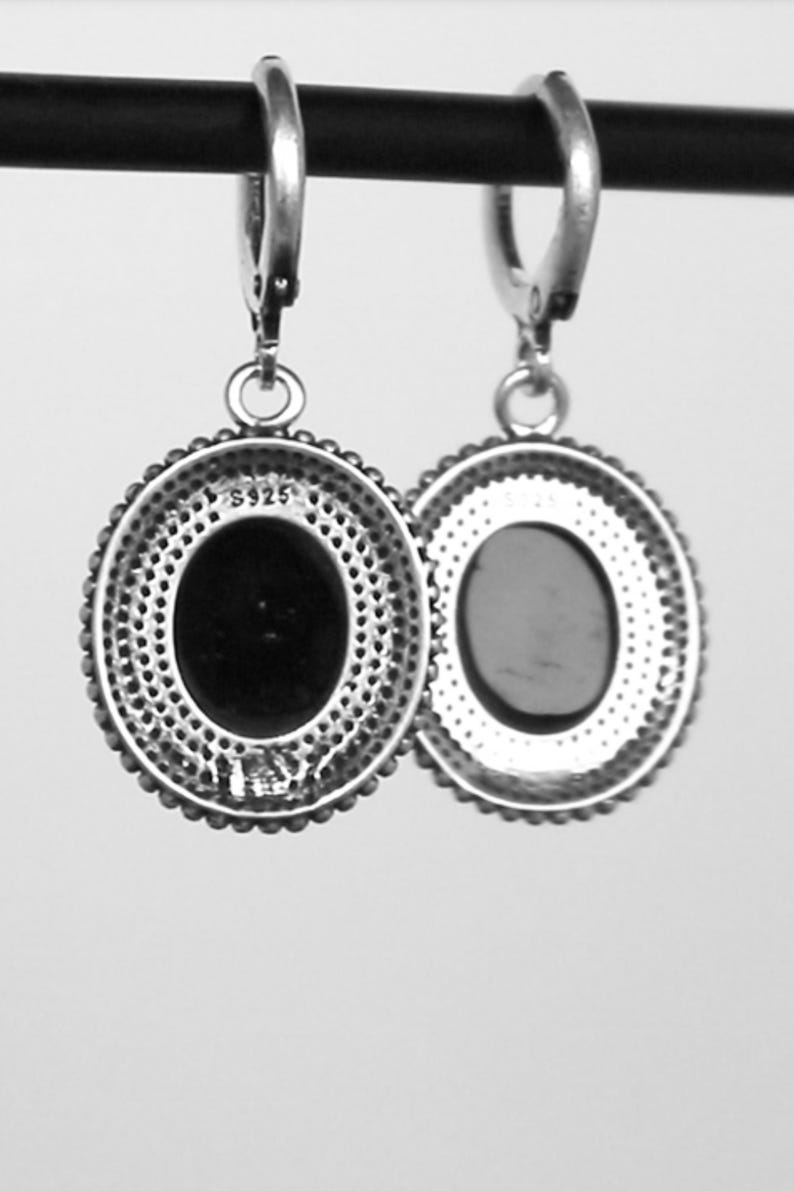May include: A pair of silver-coloured oval drop earrings. Each earring has a dark oval centre stone, a beaded border, and a decorative outer edge. The earrings are suspended from a simple hoop.