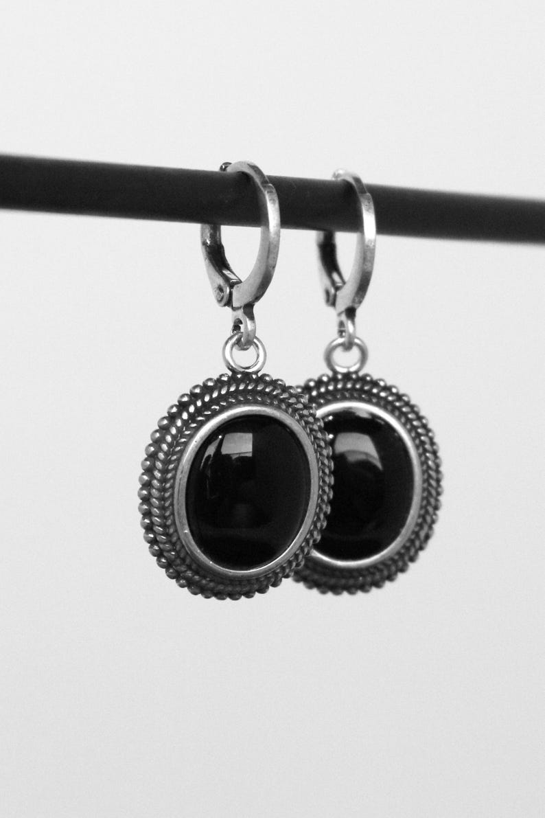May include: A pair of silver earrings featuring a black, oval gemstone. The earrings have a beaded edge and a lever-back clasp. The jewellery is presented against a plain white backdrop.