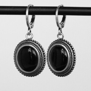 May include: A pair of silver earrings featuring a black oval gemstone. The earrings have a decorative, rope-style border and a lever-back clasp. The earrings are shown against a plain backdrop.