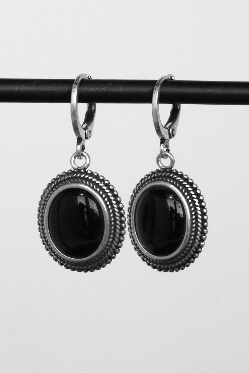 May include: A pair of silver earrings featuring a black oval gemstone. The earrings have a decorative, rope-style border and a lever-back clasp. The earrings are shown against a plain backdrop.
