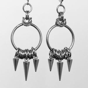 May include: A pair of silver hoop earrings. Each earring has a large circular hoop, a coil of smaller rings, and three dangling, conical spikes. The earrings are suspended from a black bar against a white backdrop.