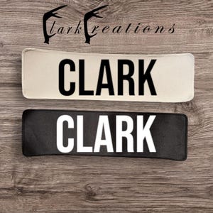 Peut inclure: Deux écussons rectangulaires, l'un crème avec du texte noir et l'autre noir avec du texte blanc, affichant tous deux le nom "CLARK". Les écussons sont posés sur un fond en bois, avec le texte "Clark Creations" au-dessus.