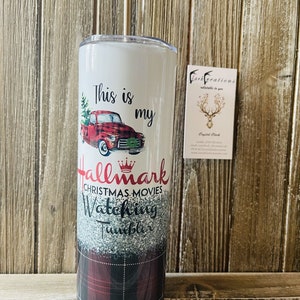 May include: A white tumbler with a red truck and Christmas tree design. The text "This is my Hallmark Christmas Movies Watching Tumbler" is printed on the tumbler. The tumbler has a plaid pattern on the bottom half.