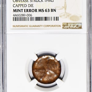 May include: A Lincoln Wheat 1C coin, graded MS 63 BN, encased in a protective holder. The coin features an obverse struck thru capped die error. The holder includes the text "Big Liberty" and details from the Numismatic Guaranty Corporation.