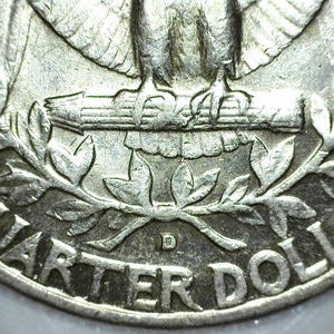 May include: A silver-colored United States quarter dollar coin featuring a detailed relief of an eagle perched on a branch. The text QUARTER DOLLAR is embossed along the bottom edge, with a small D mint mark visible above the word QUARTER.