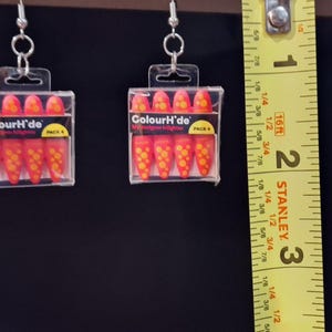 May include: A pair of dangle earrings featuring miniature clear plastic cases containing four orange and yellow highlighter markers labeled ColourH'de. A yellow Stanley measuring tape is positioned next to the earrings, showing a length of 1 inch.