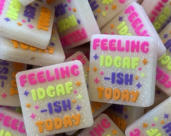 Könnte beinhalten: Ein Set aus weißen, glitzernden quadratischen Schlüsselanhängern mit dem Spruch "Feeling IDGAF-ish Today" in verschiedenen Farben. Die Schlüsselanhänger sind aus einem weichen, flexiblen Material gefertigt und haben ein lustiges, skurriles Design.