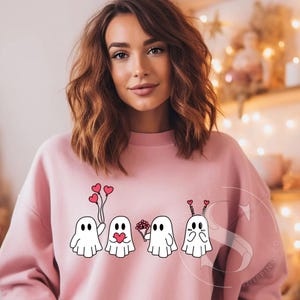 May include: Pink sweatshirt featuring a design of four ghosts holding hearts and flowers. The ghosts are white with black eyes and mouths. The hearts are red and the flowers are pink.