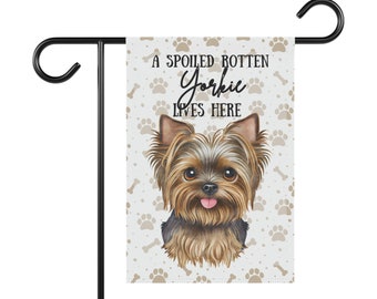 May include: A white garden flag featuring a brown and black Yorkshire Terrier dog illustration. The flag has a white background with brown paw prints and bone patterns. The text on the flag reads "A Spoiled Rotten Yorkie Lives Here".
