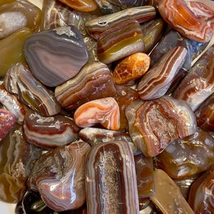 Puede incluir: Una colección de piedras de ágata en varios tonos de marrón, rojo y blanco. Las piedras son lisas y pulidas, con intrincados patrones de bandas.