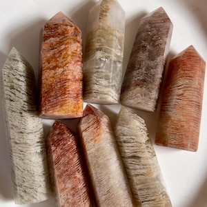 May include: A collection of eight quartz crystal towers in various shades of orange, brown, and cream. The crystals are polished and have a pointed top. The crystals are arranged on a white surface.