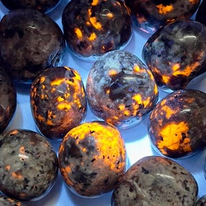 May include: A collection of polished, rounded stones with a speckled appearance. The stones have a dark, mottled base color with vibrant orange and yellow glowing inclusions, creating a striking contrast. The stones are arranged closely together.