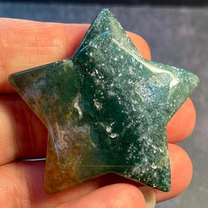 May include: A polished star-shaped gemstone with a moss agate pattern. The stone features a mix of deep green, white, and golden-brown hues. The star is held in a hand, showcasing its natural beauty and unique color variations.