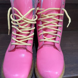 Puede incluir: Un par de botas de cordones de charol rosa con cordones amarillos. Las botas tienen una suela negra y un detalle de costura blanca.
