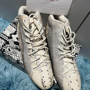 May include: A pair of white trainers with a snakeskin pattern. The trainers have white laces and a white sole. The text "To Clean Wipe With Damp Cloth Do Not Machine Wash Design Patent Pending" is on a white card behind the trainers.