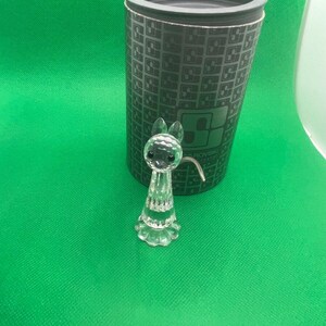 May include: A clear crystal bunny figurine with a long, slender body and a flared base. The figurine is positioned in front of a dark green cylindrical container with a geometric pattern. The background is a solid green surface.