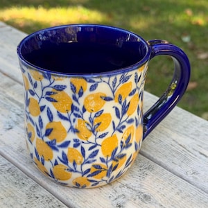 May include: A ceramic mug with a deep blue interior and handle, featuring a cream-colored exterior decorated with a repeating pattern of yellow lemons and dark blue leafy branches. The mug has a rounded shape and a smooth, glossy finish throughout.