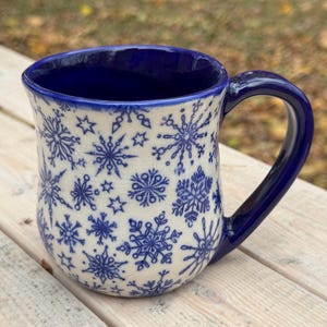May include: A ceramic mug with a blue interior and handle. The exterior is off-white with a pattern of blue snowflakes and stars. The mug is sitting on a light-colored wooden surface.