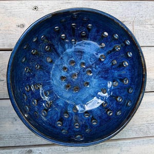 May include: A round ceramic colander bowl featuring a deep cobalt blue glaze with lighter blue swirling variations. The bowl has a smooth, glossy finish and is perforated with numerous evenly spaced circular drainage holes throughout the basin.