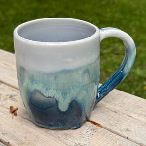 May include: A ceramic mug featuring a smooth, light gray upper section that transitions into a reactive glaze with shades of teal, turquoise, and deep navy blue. The mug has a rounded body, a wide opening, and a curved handle with a matching blue finish.