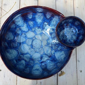 Op de afbeelding: Een handgemaakte keramische serveerschaal met een aangehechte dipschaal. De schaal heeft een diepblauwe glazuur met een textuur, bubbelachtig patroon. De rand is een donkerdere tint blauw.
