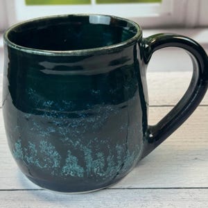 Puede incluir: Una taza de cerámica hecha a mano con un esmalte verde oscuro. La taza tiene una forma redondeada con un asa grande. El esmalte tiene un aspecto texturizado y moteado con acentos verde claro cerca de la base.