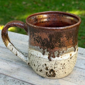 May include: A ceramic mug featuring a two-tone glaze with a dark metallic brown upper section that transitions into a speckled cream base. The mug has a curved handle and a wide rim, showcasing a textured finish where the brown glaze drips over the white.