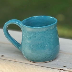 Puede incluir: Taza de cerámica turquesa hecha a mano. Presenta un cuerpo redondeado, un asa curva y una superficie texturizada. Ideal para bebidas frías o calientes.