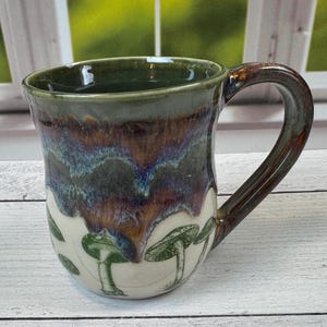 May include: A handmade ceramic mug with a dark green rim and handle. The mug features a unique glaze with shades of blue, brown, and green. The lower portion of the mug is white with green mushroom illustrations. A functional and decorative piece.