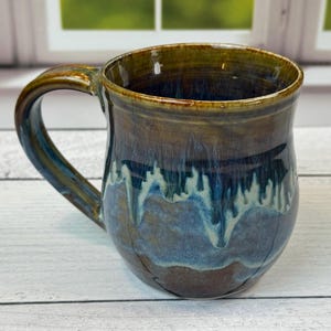 Könnte beinhalten: Eine handgefertigte Keramiktasse mit einer einzigartigen Glasur. Die Tasse hat eine dunkelbraune Außenseite, die sich zum Boden hin in Blau und Weiß verwandelt. Der Griff ist in einem dunkleren Braunton gehalten. Ein schönes Stück für Kaffee oder Tee.