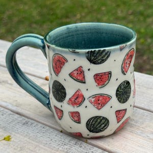 Pode incluir: Uma caneca de cerâmica com alça e interior azul-petróleo. O exterior é branco com um padrão de fatias e melancias inteiras em vermelho e verde. A caneca está sobre uma superfície de madeira clara.