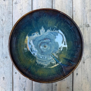 Peut inclure: Un bol en céramique rond avec un vernis bleu profond et vert à la finition brillante. L'intérieur présente un motif tourbillonnant de bleu clair et de blanc rayonnant depuis le centre, passant par des tons bleu foncé et vert terreux.
