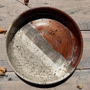May include: A round ceramic bowl with a high rim, featuring a multi-toned glaze. The interior displays a transition from a speckled cream and grey base to a textured, bubbly grey-brown band, ending in a smooth, deep reddish-brown glazed section.