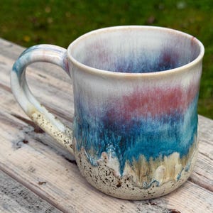 May include: A ceramic mug featuring a reactive glaze with a gradient of cream, deep blue, and reddish-purple tones. The base of the mug has a speckled texture, while the upper portion displays a smooth, blended finish. It includes a curved handle.