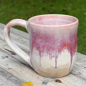 Peut inclure: Une tasse en céramique dotée d'une anse arrondie et d'un corps cylindrique. L'extérieur présente un émail réactif avec un dégradé passant d'un beige crème doux à la base à un rose saturé et vibrant qui coule vers le bas.