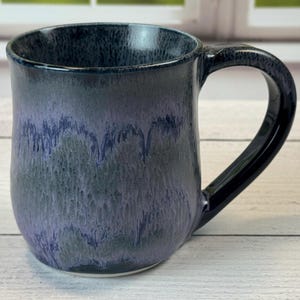 Puede incluir: Una taza de cerámica con un asa azul oscuro y un cuerpo que se desvanece del azul oscuro al azul claro y gris. La taza tiene un acabado brillante y una forma redondeada, perfecta para café o té.