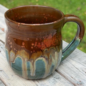 May include: A ceramic mug featuring a glossy glaze with a deep brown upper section that transitions into a mottled reddish-brown and a teal-green base. The glaze exhibits a distinct drip pattern where the colors overlap, and it has a curved handle.