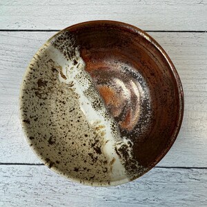 May include: A handmade ceramic bowl with a unique design. The bowl features a split design with a white and speckled tan side, and a rich brown side. The interior has a glossy finish, and the bowl is perfect for serving food or as a decorative piece.