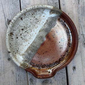 May include: A round ceramic spoon rest featuring a dual-tone glaze. One side is a speckled off-white with dark brown flecks, transitioning into a textured, bubbly grey band, and finishing with a smooth, glossy deep reddish-brown glaze on the other side.