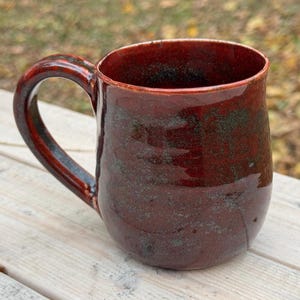 May include: A handmade ceramic mug with a deep red glaze and a dark handle. The mug has a glossy finish with subtle green and black speckles. The interior is also red, and the mug sits on a light-colored wooden surface.