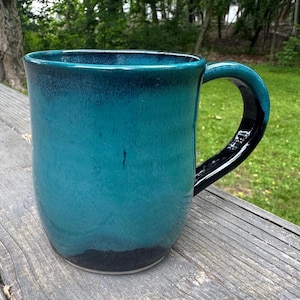 Pode incluir: Uma caneca de cerâmica com esmalte turquesa e preto. A caneca tem um corpo arredondado e uma alça grande e curva. A cor turquesa desvanece-se para preto na parte superior e inferior. A caneca está sobre uma superfície de madeira.