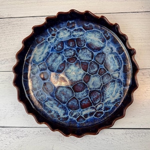 Puede incluir: Un plato redondo de cerámica con un borde marrón oscuro y ondulado. El interior presenta un patrón azul y marrón que se asemeja a un mosaico o estructura celular. La superficie del plato es brillante.
