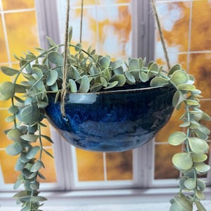 May include: A hanging planter featuring a deep blue ceramic bowl filled with faux eucalyptus. The bowl is suspended by twine. This decorative piece adds a touch of nature to any interior space, with a diameter of approximately 20 cm.