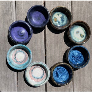 May include: Seven small ceramic bowls in various shades of blue, green, and white with swirling patterns. The bowls are arranged in a circle on a wooden surface.