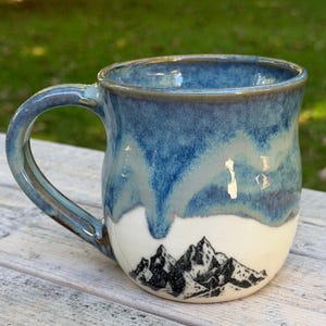 May include: A ceramic mug featuring a glossy blue glaze that drips down from the rim over a white base. The lower portion of the mug displays a detailed black ink illustration of a mountain range, complete with jagged peaks and textured slopes.