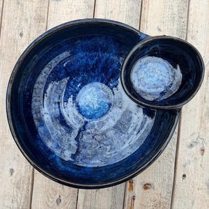 Op de afbeelding: Een handgemaakte, donkerblauwe keramische serveerschaal met een bevestigde kleinere kom. De schaal heeft een glanzende afwerking met een marmereffect, met tinten blauw en wit. Het ontwerp is ideaal voor het serveren van snacks of dips.