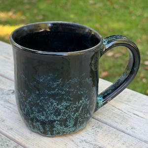 May include: A ceramic mug with a glossy black finish and a textured, mottled teal-green glaze pattern concentrated toward the base. The mug features a wide, curved handle and a smooth, rounded rim, showcasing a deep, dark interior surface.